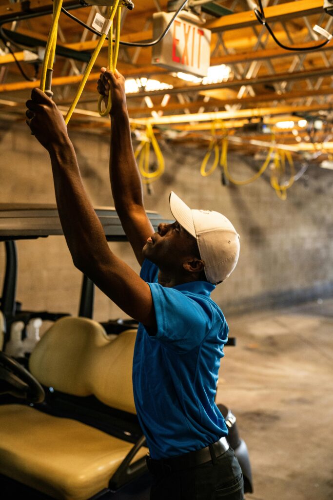 A man wearing a cap works on wiring above a golf cart in a garage.
